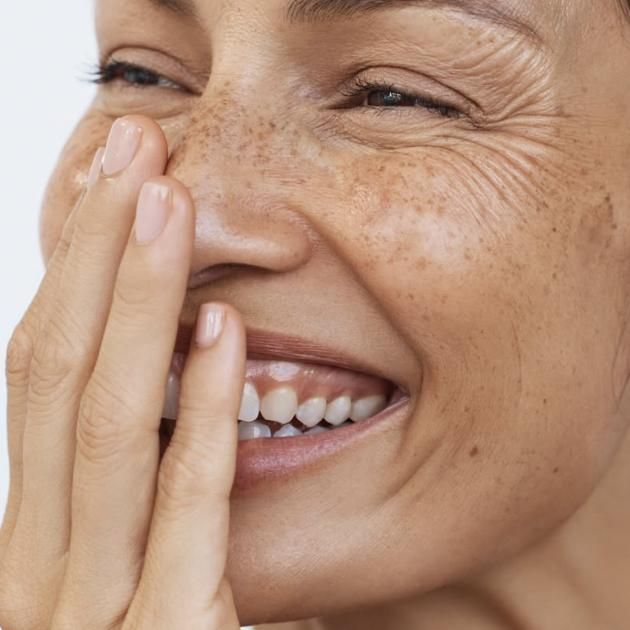 A woman with natural freckles and age spots looking into a mirror in a bright, modern Sydney clinic setting, representing healthy skin aging.