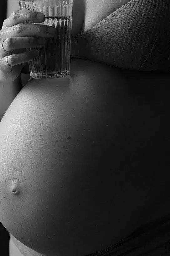 A close-up black and white image of a pregnant woman holding a glass of water, symbolizing the importance of hydration in a pregnancy-safe skincare routine by Body Sculpting Clinics Sydney.
