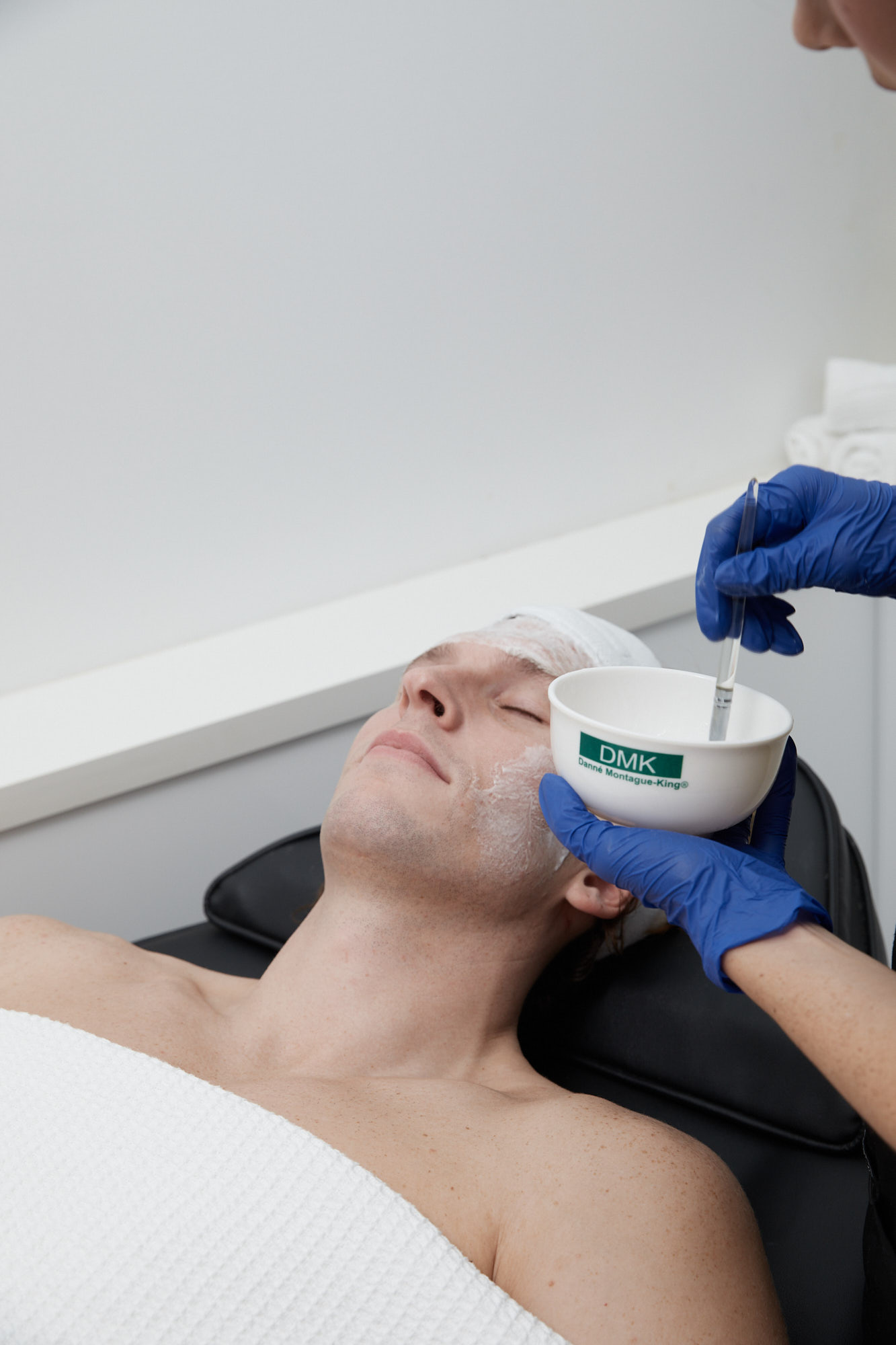 A man lying down in a clinic setting while receiving a professional DMK enzyme therapy skin treatment on his face.