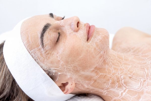 Close-up profile of a woman lying down with a hardened, tight DMK enzyme masque applied across her face, neck, and décolletage.