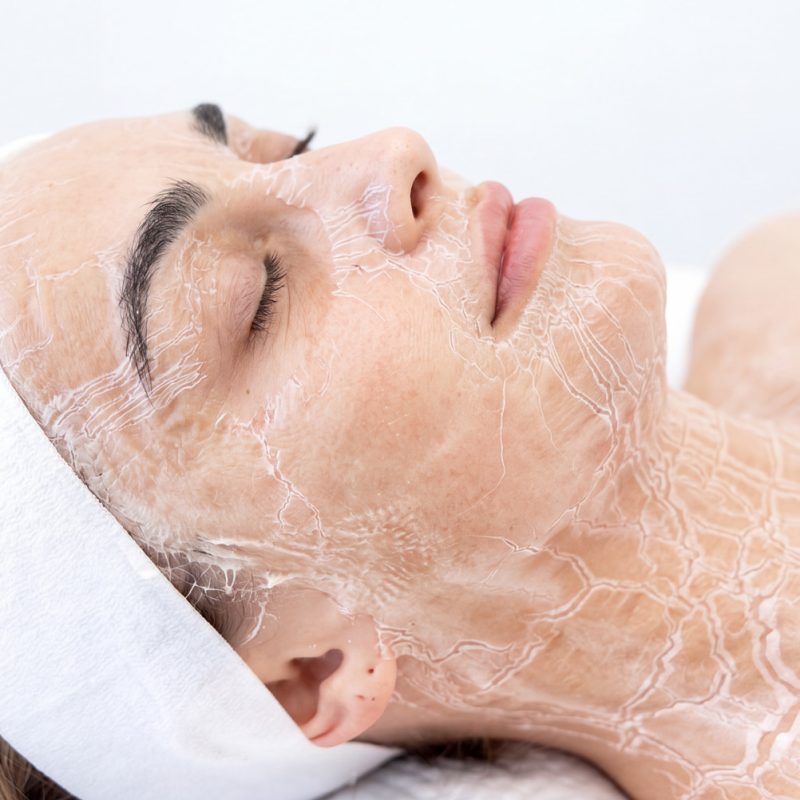 Close-up profile of a woman lying down with a hardened, tight DMK enzyme masque applied across her face, neck, and décolletage.