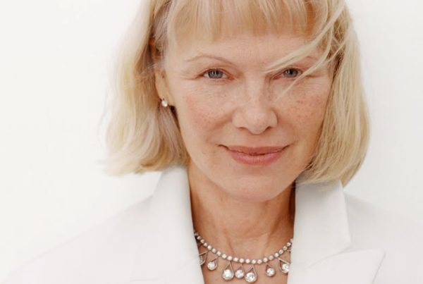 Close-up portrait of a mature woman with blonde hair, soft freckles, and a subtle smile in a white blazer, highlighting natural skin texture.
