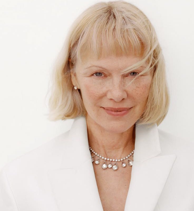 Close-up portrait of a mature woman with blonde hair, soft freckles, and a subtle smile in a white blazer, highlighting natural skin texture.