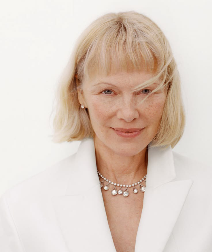 Close-up portrait of a mature woman with blonde hair, soft freckles, and a subtle smile in a white blazer, highlighting natural skin texture.